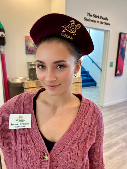 Emma museum children’s curator wearing a maroon Beret with 'OMAM' Museum Turtle Logo. She is inside the Ormond Memorial Arts and Gardens Museum.

