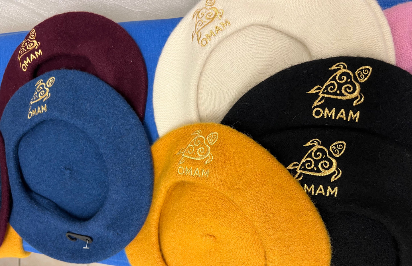 Collection of berets in various colors with 'OMAM' branding gold embroidered turtle logo.